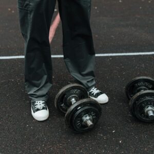 Individual in sneakers and trousers stands near dumbbells on an outdoor track, ready for fitness training.