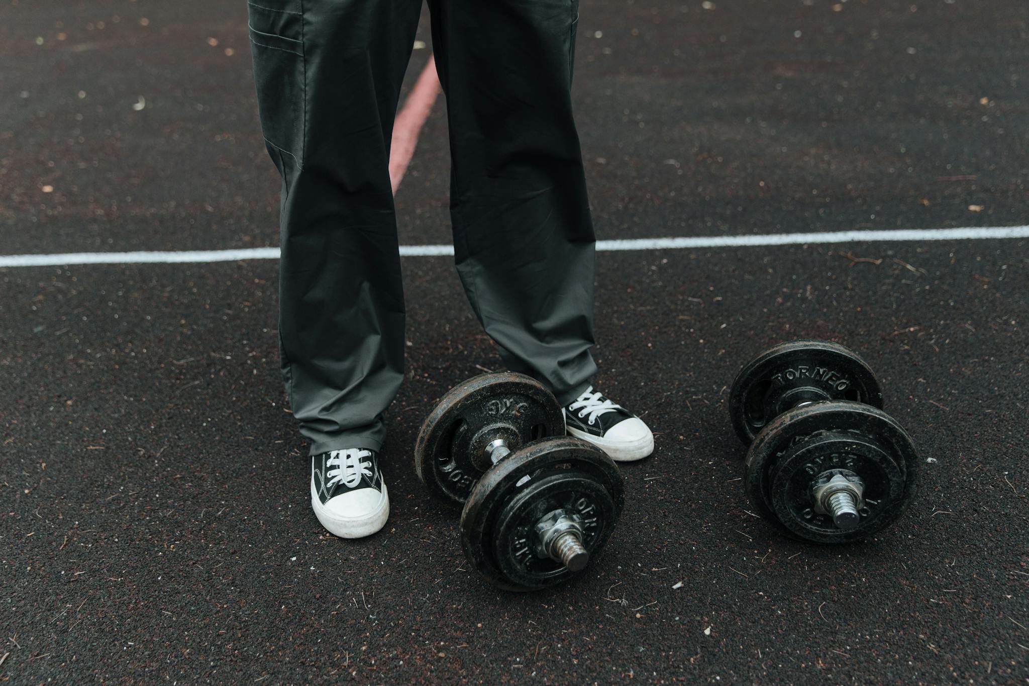 Individual in sneakers and trousers stands near dumbbells on an outdoor track, ready for fitness training.