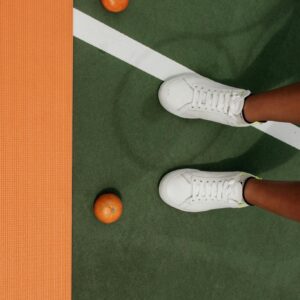 White sneakers on a tennis court with scattered oranges, highlighting sports and lifestyle theme.