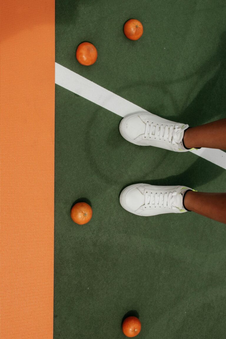 White sneakers on a tennis court with scattered oranges, highlighting sports and lifestyle theme.