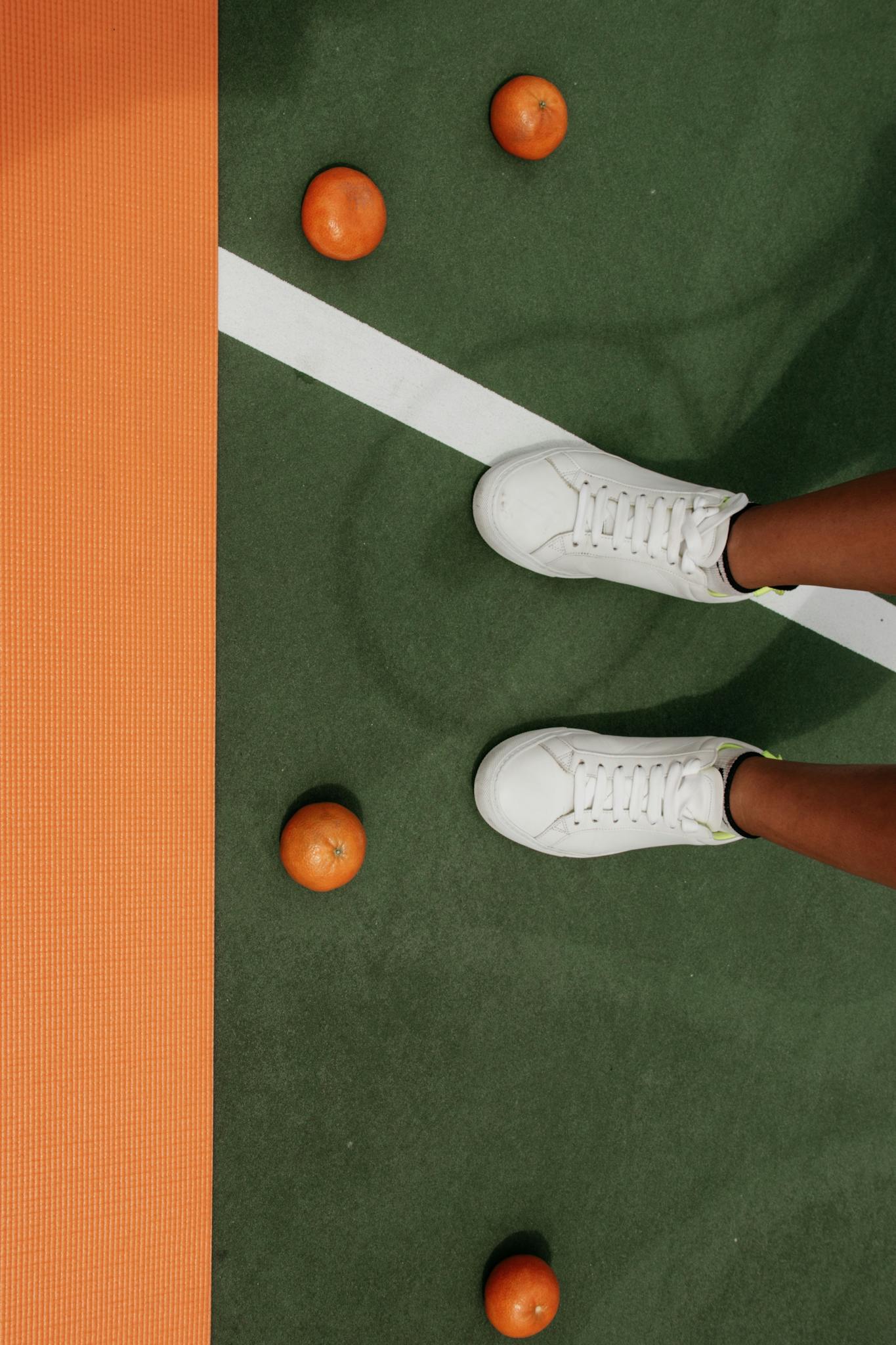 White sneakers on a tennis court with scattered oranges, highlighting sports and lifestyle theme.
